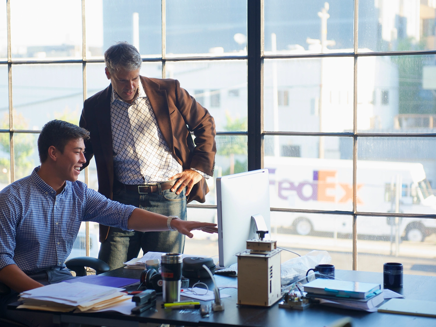 Two men in an office discussing something on a computer, with a FedEx truck visible through the window