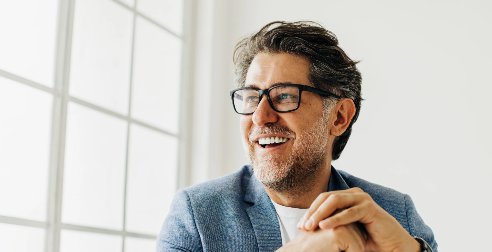 Smiling man in glasses and a blue blazer sitting by a large window