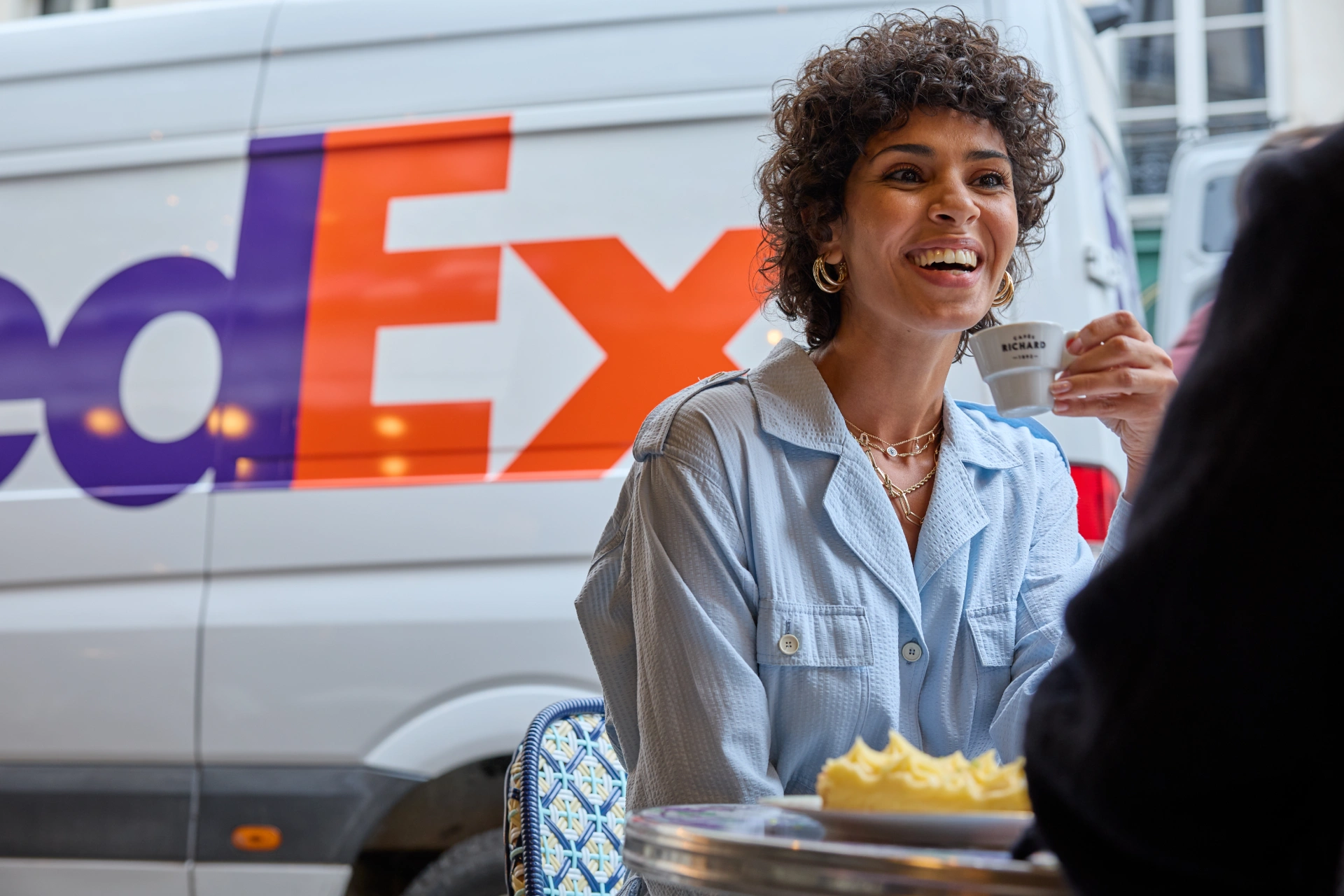 Smiling woman drinking espresso at an outdoor café, with a FedEx van in the background