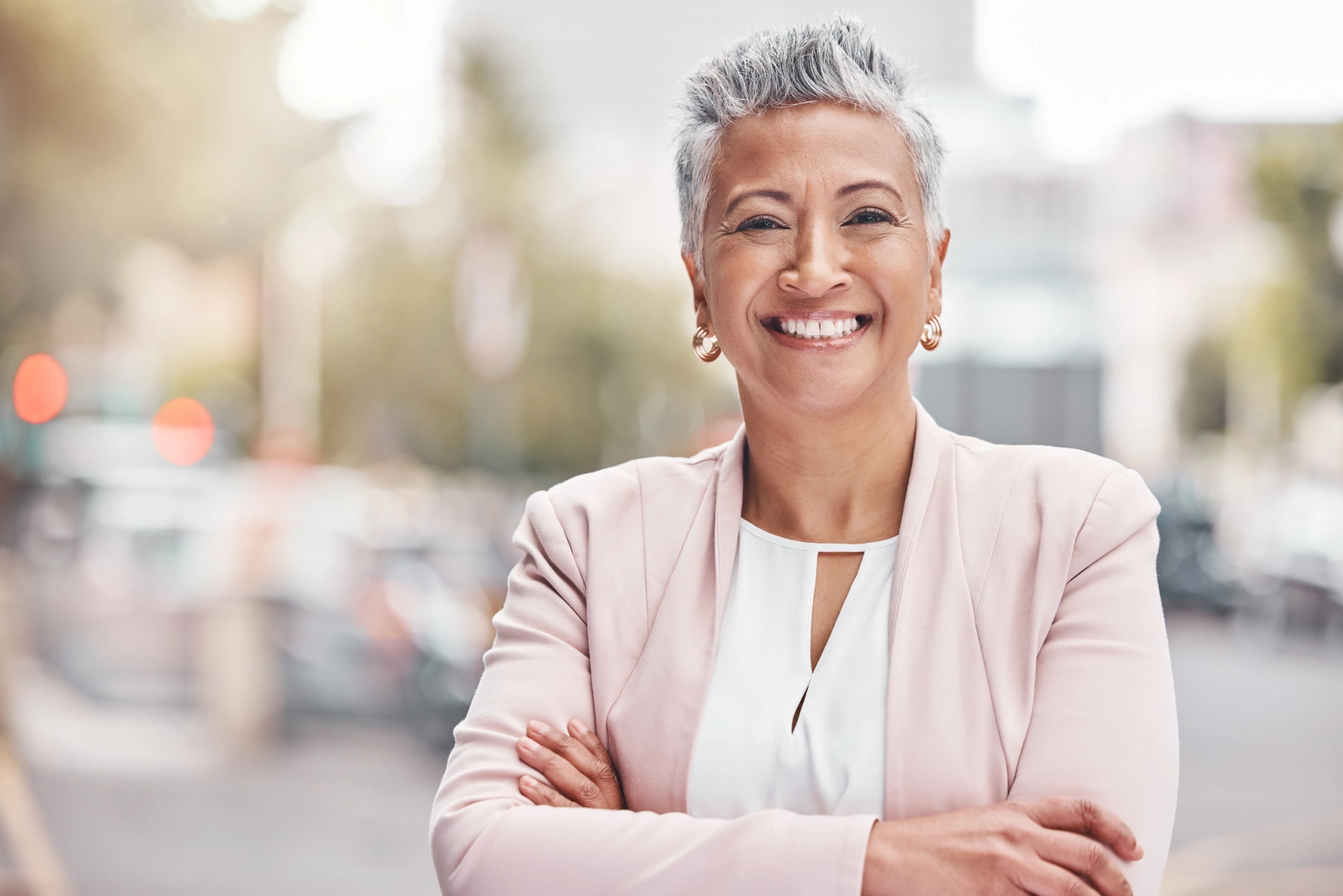 Confident mature woman with short gray hair smiling and standing with arms crossed, wearing a light pink blazer and white blouse, in an urban outdoor setting