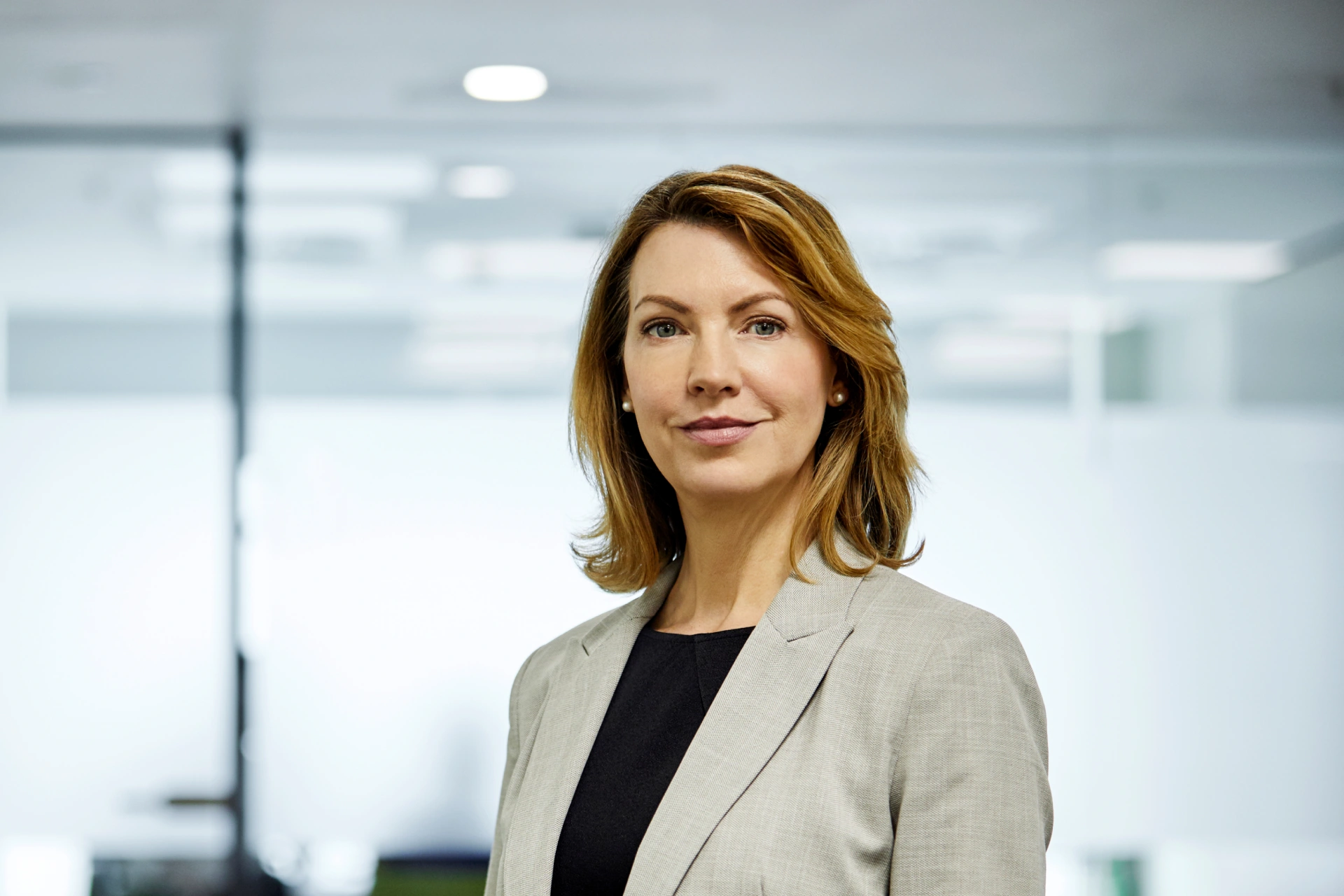 Confident businesswoman with shoulder-length light brown hair, wearing a light grey blazer and black top, standing in a modern office setting